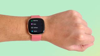 En rosa Fitbit Versa 4 runt en persons handled som hålls upp framför en mintgrön bakgrund.