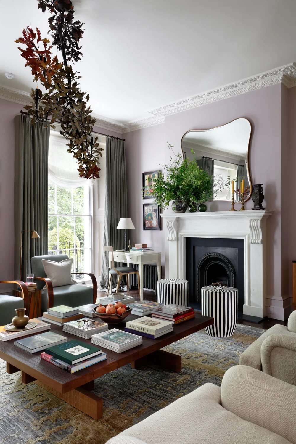 A layered living room interior with pale lilac walls, stuccoed detailing, wood and upholstered furniture, striped black and white stools, and lots of books on a coffee table along with art and plants.