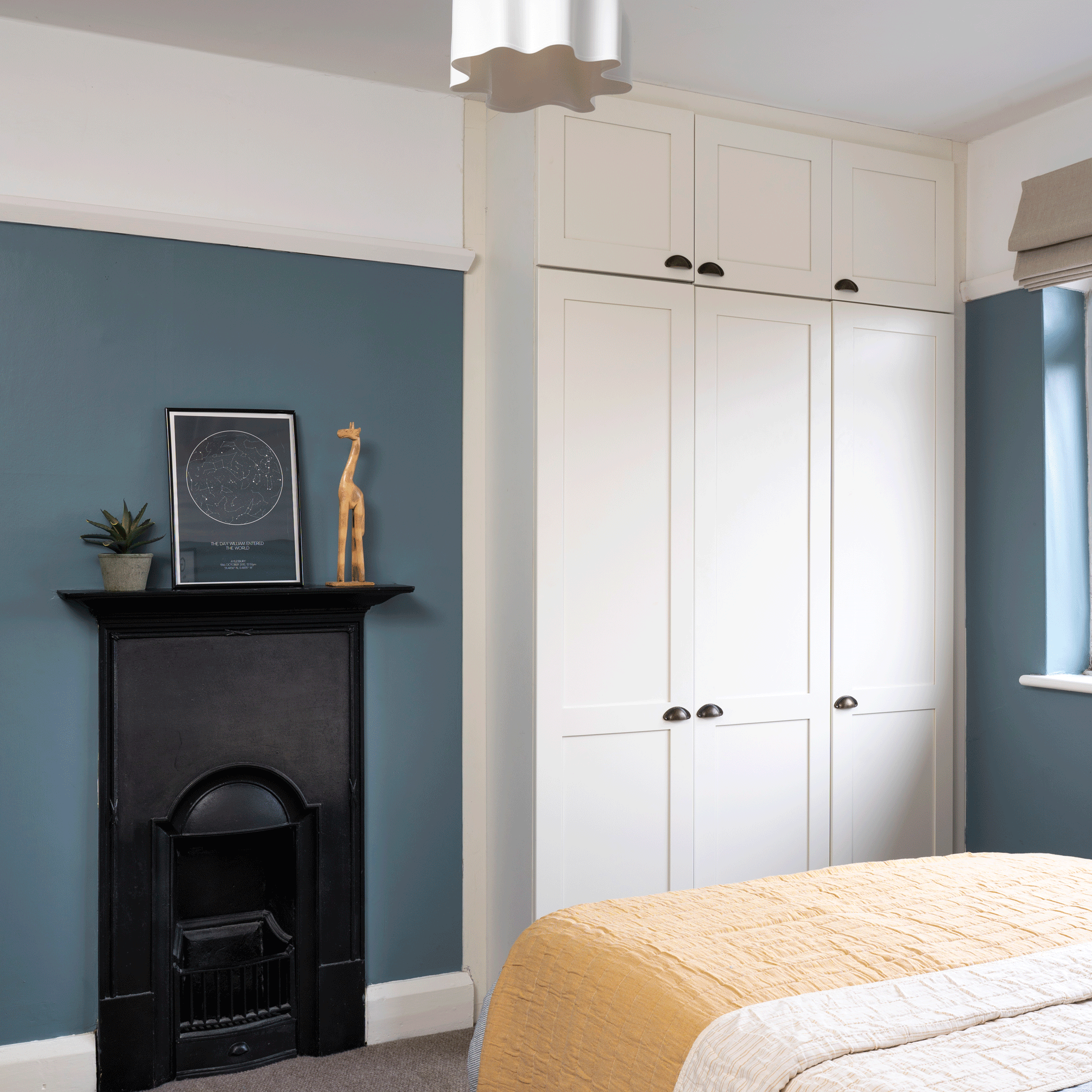 a bedroom with dark blue painted walls white built in wardrobes a black traditional cast-iron fireplace and a bed with a yellow bedspread