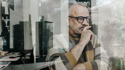 An older man looks thoughtful as he looks out his office window, a skyline reflected in the glass.