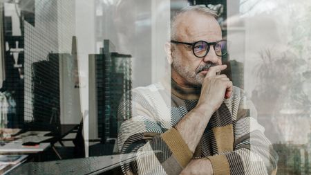 An older man looks thoughtful as he looks out his office window, a skyline reflected in the glass.