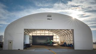 A jet with pale green panel coverings and sleek, sharp edges and angles sits underneath the canopy of a hanger, the bright sun peaking just above the structure's right side, against a cloudy blue sky. 