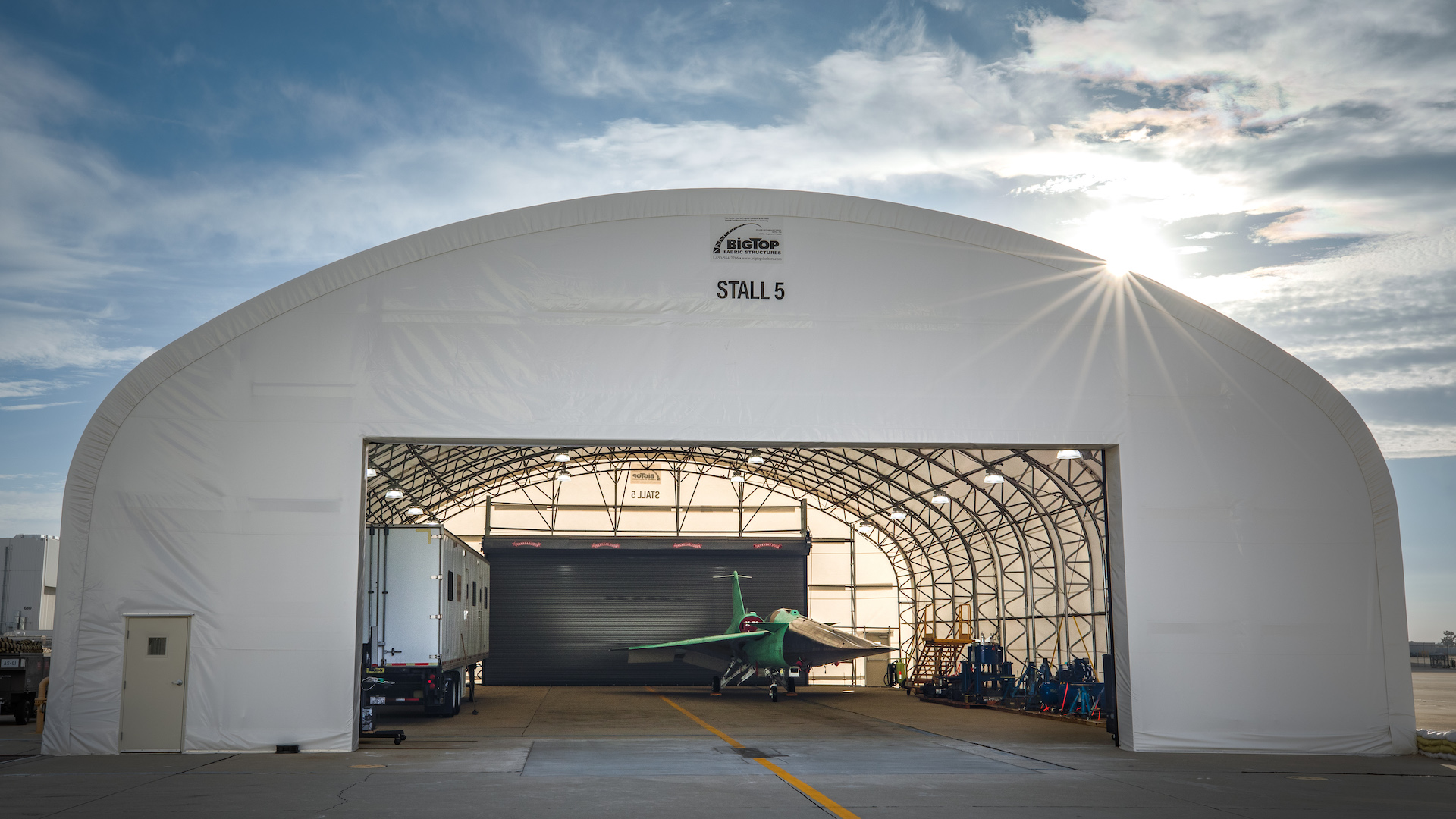 A jet with pale green panel coverings and sleek, sharp edges and angles sits underneath the canopy of a hanger, the bright sun peaking just above the structure's right side, against a cloudy blue sky.