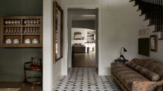 A tiled entryway with a patterned couch by the stairs, a path to the living room, and a white wall with a mirror separating the dining room with floating storage displaying china