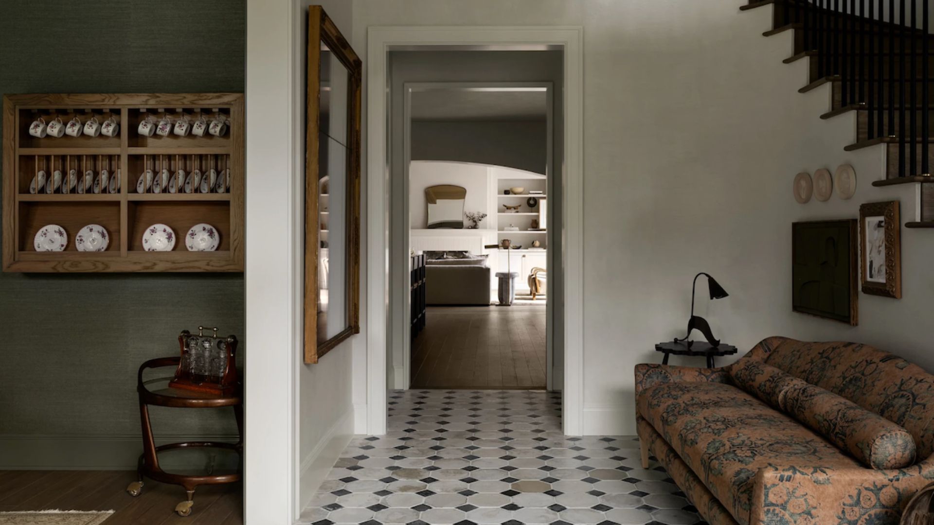 A tiled entryway with a patterned couch by the stairs, a path to the living room, and a white wall with a mirror separating the dining room with floating storage displaying china
