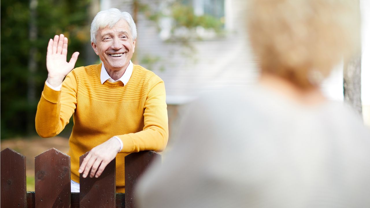 An older man waves hello to his neighbor over his fence.