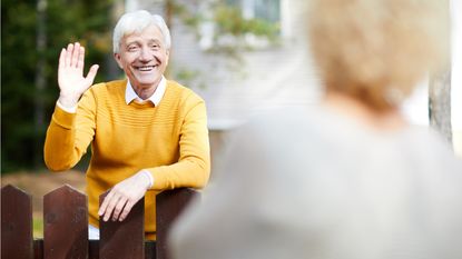 An older man waves hello to his neighbor over his fence.