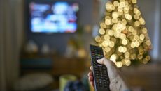 Hand on remote control pointing at TV, with a lit Christmas tree beside the TV