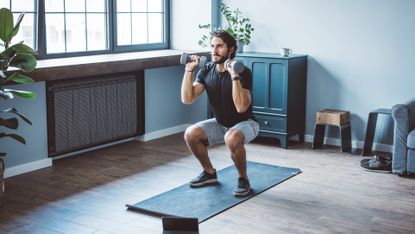 A man performs a dumbbell squat in a living room on a yoga mat. His knees are bent, thighs parallel with the floor, and he holds the dumbbells near his shoulders. Behind him we see a cupboard, a plant and a stool. 