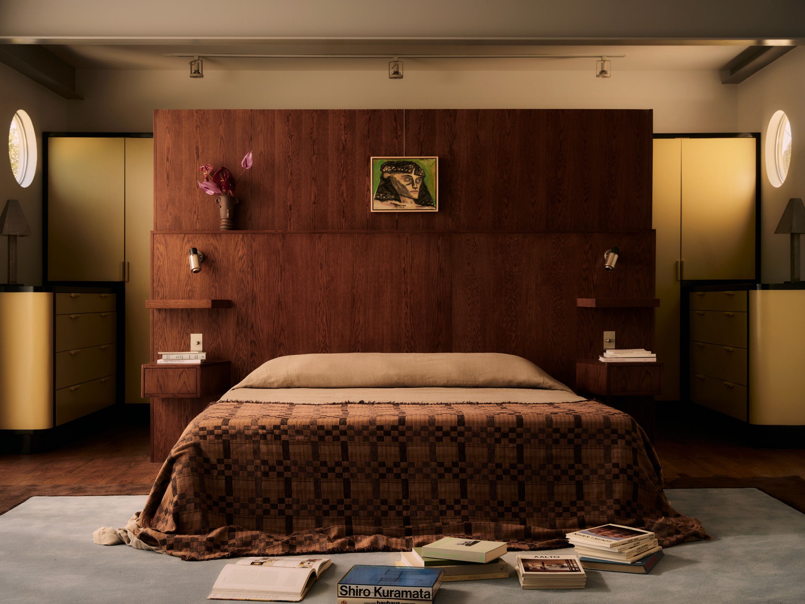 A modern bedroom with a large wooden headboard with built-in nightstands and shelving, with portholes on either side of the bed which is draped with a patterned bedspread that puddles on the floor