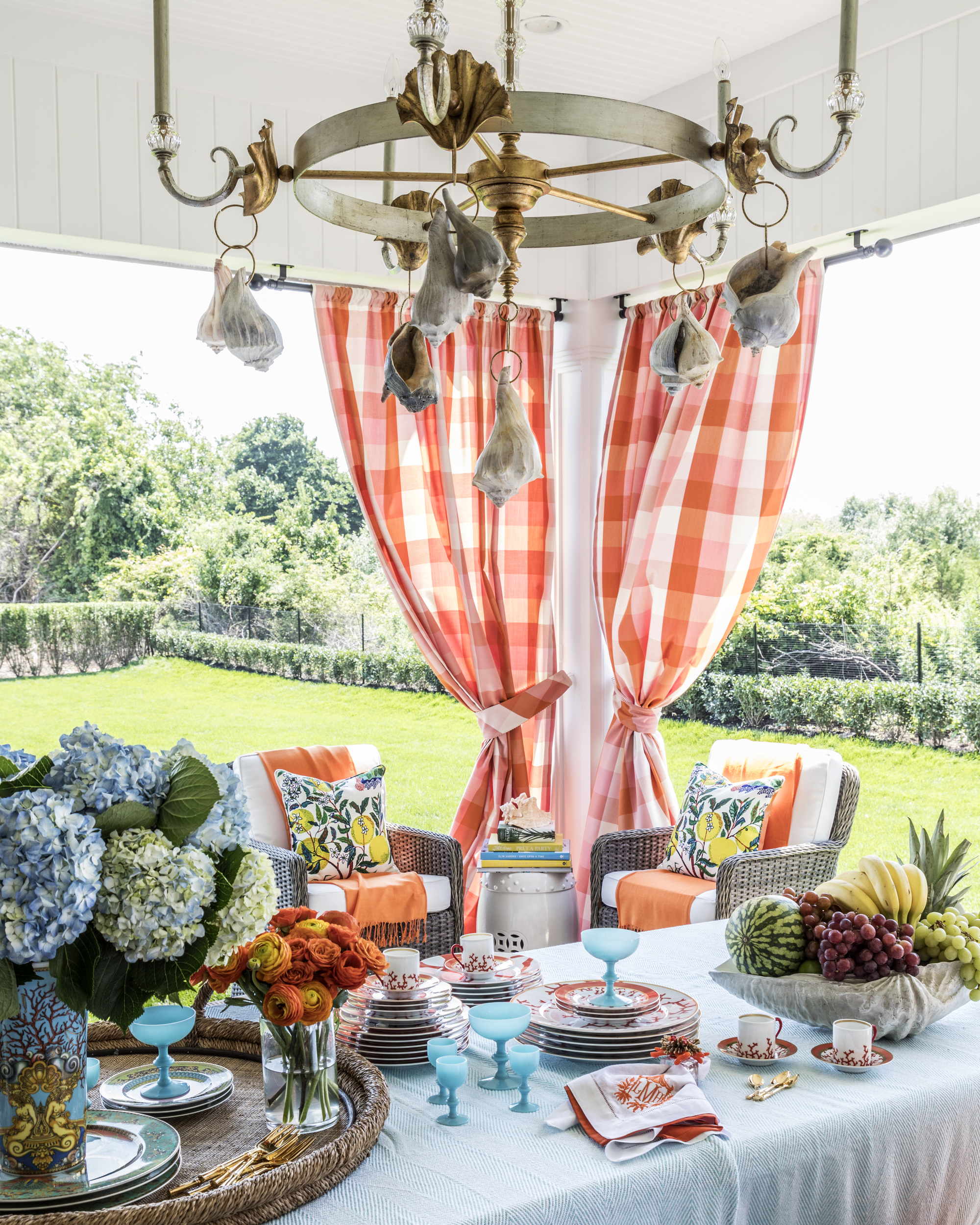 A pergola with a table laid with dinner plates, vases, and fruit, and with red and white checkered curtains hanging up