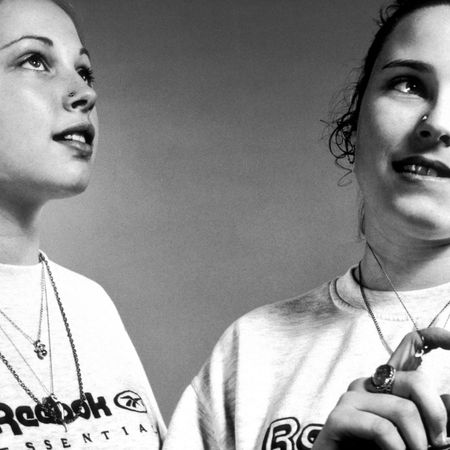 Black-and-white photograph of two young women wearing white Reebok T-shirts, layered necklaces and hoop earrings, looking off-camera.