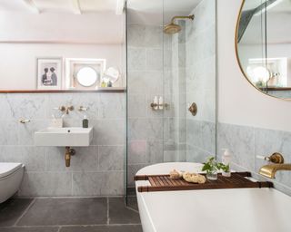 Bathroom with white bath, glass shower cubicle, basin and toilet, grey wall tiles and pale grey walls.