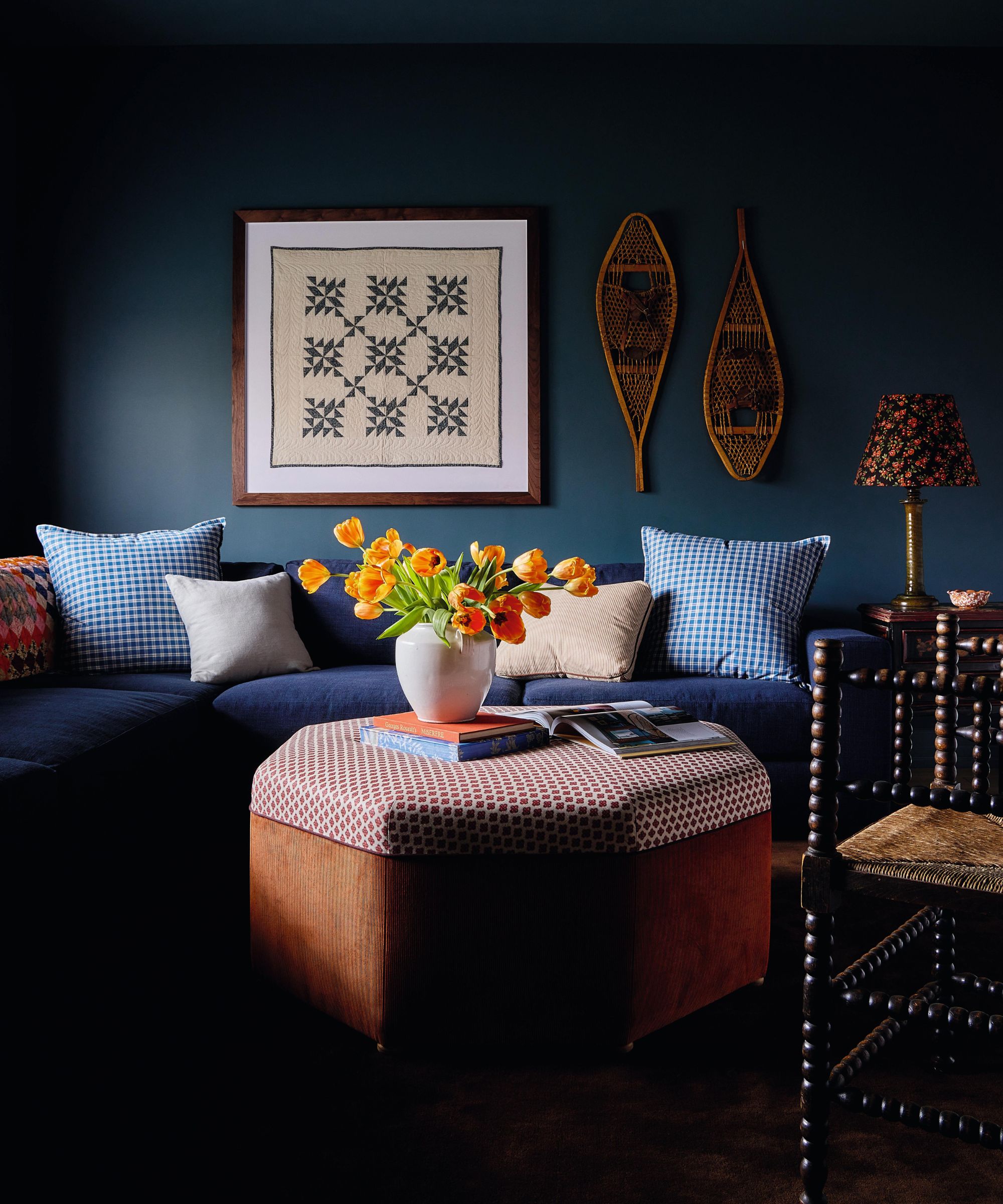 a media room painted navy with a navy couch and plaid pillows, a patchwork piece of art, rattan decor, ottoman and bobbin chair