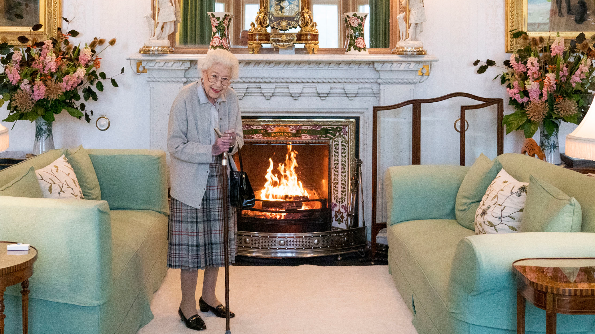 Queen Elizabeth wearing a plaid skirt standing in front of a fireplace and green sofa