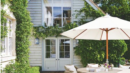 A sunny patio with a circular table with white umbrella, in front of a white panelled house with large windows and doors, and green climbing plants all over it.