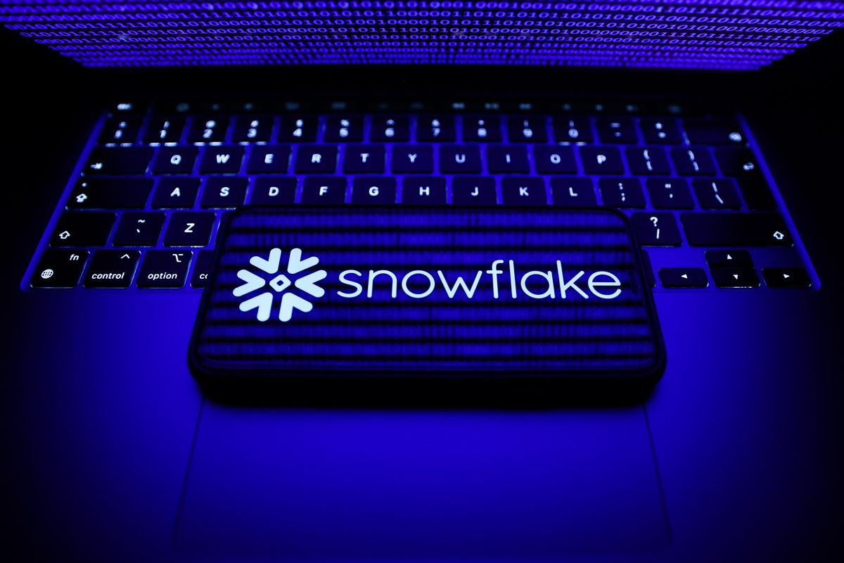 Snowflake company logo appearing on a smartphon that&#039;s laid in landscape orientation on top of a MacBook&#039;s keyboard with the computer&#039;s lid half closed, beaming a blue light onto the keyboard and phone