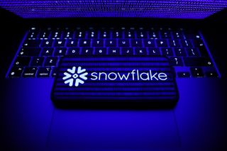 Snowflake company logo appearing on a smartphon that's laid in landscape orientation on top of a MacBook's keyboard with the computer's lid half closed, beaming a blue light onto the keyboard and phone