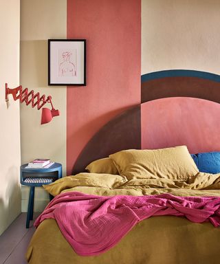 Colorful bedroom with abstract painted feature walls, mix of shapes and colors, yellow bedding with pink throw, pink wall light, rounded blue bedside table, painted purple wooden floor