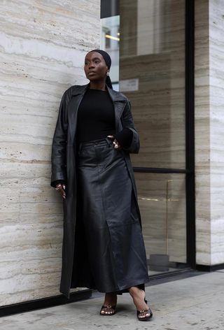 a black maxi skirt outfit showcasing a woman wearing a black stretchy headband with silver earrings, a long black leather jacket layered over a black crewneck top and black leather maxi skirt, paired with black mule sandals