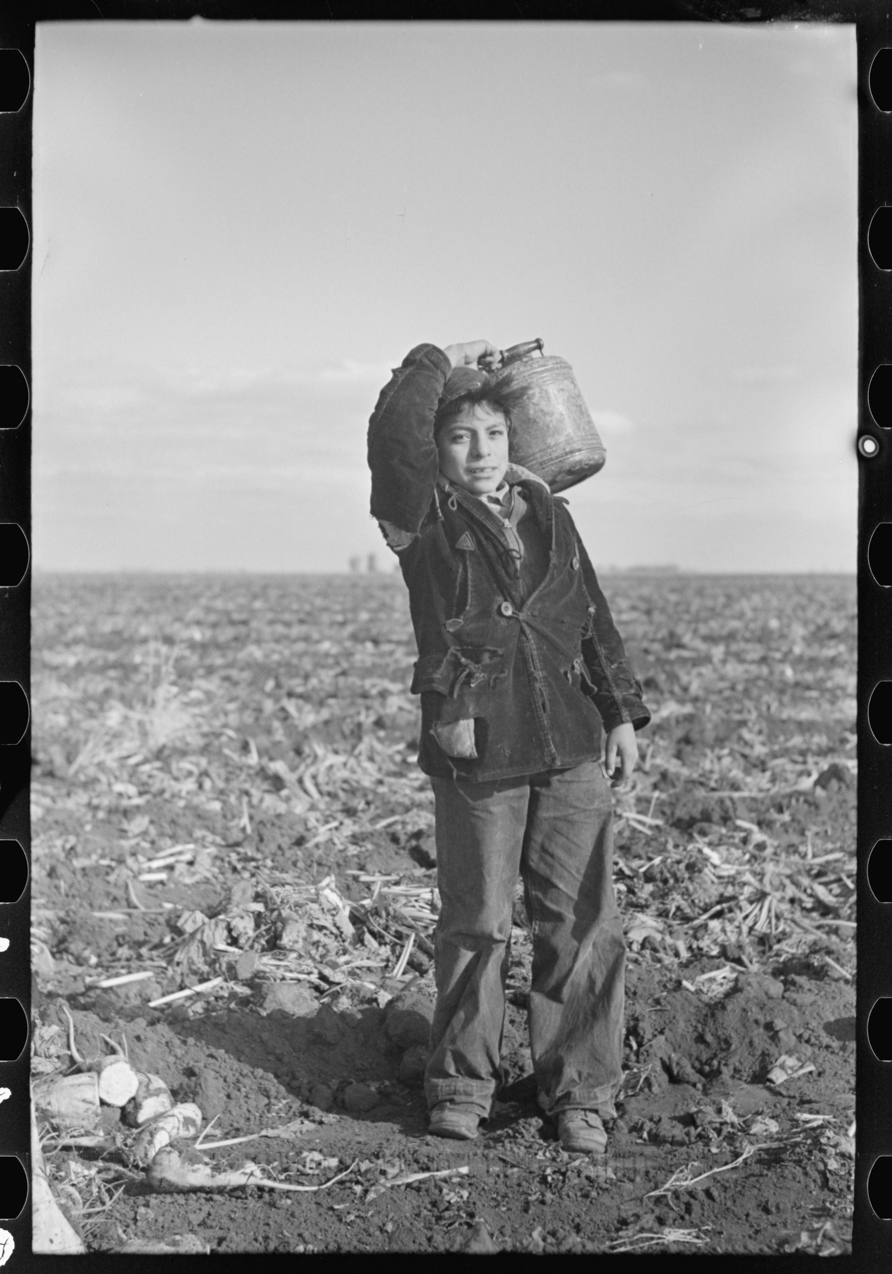 A boy in a worn corduroy jacket stands in a bare, furrowed field, balancing a large metal water can on his shoulder.