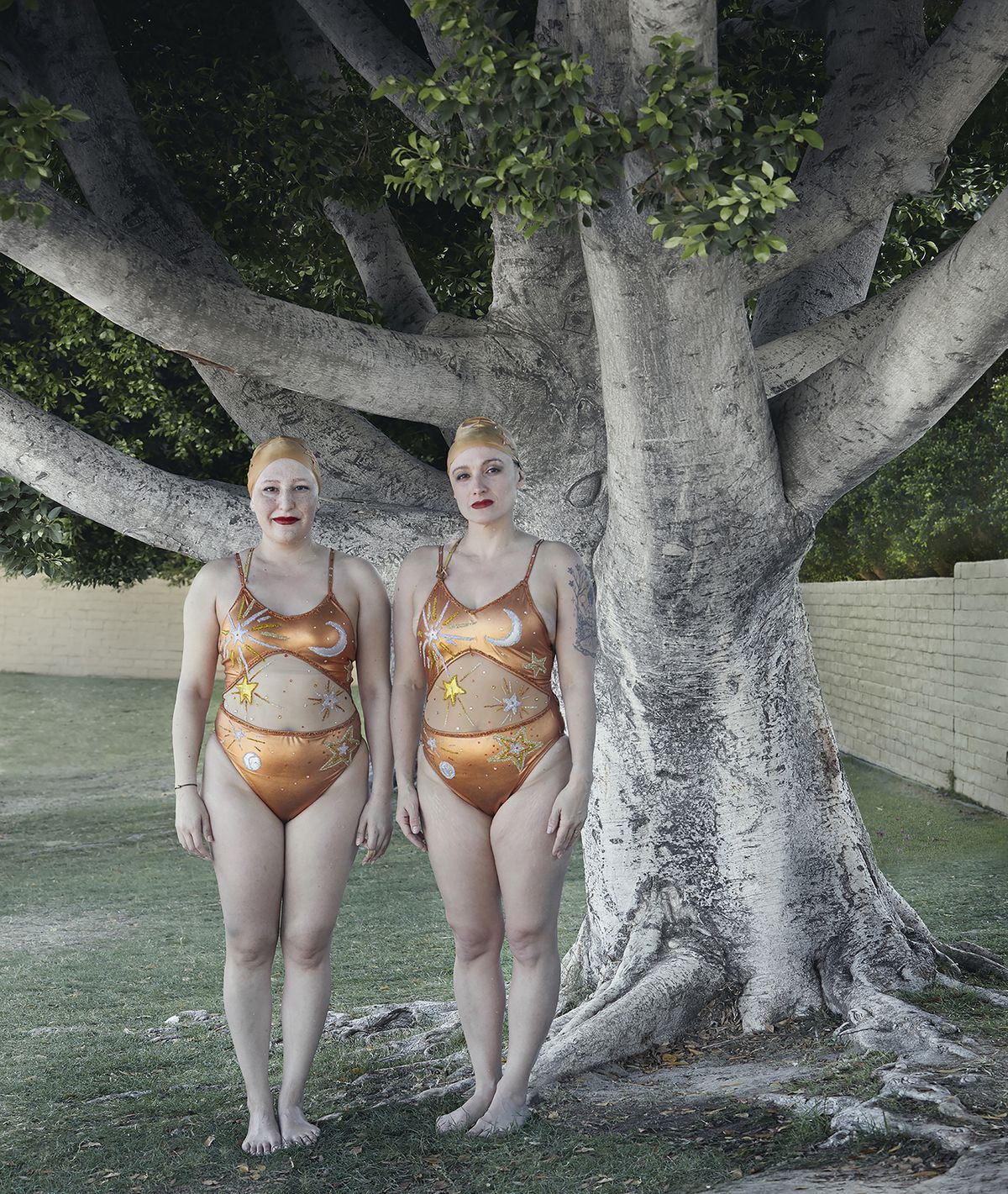 Two women in matching orange swimsuits with celestial designs stand barefoot in front of a large tree in a grassy area