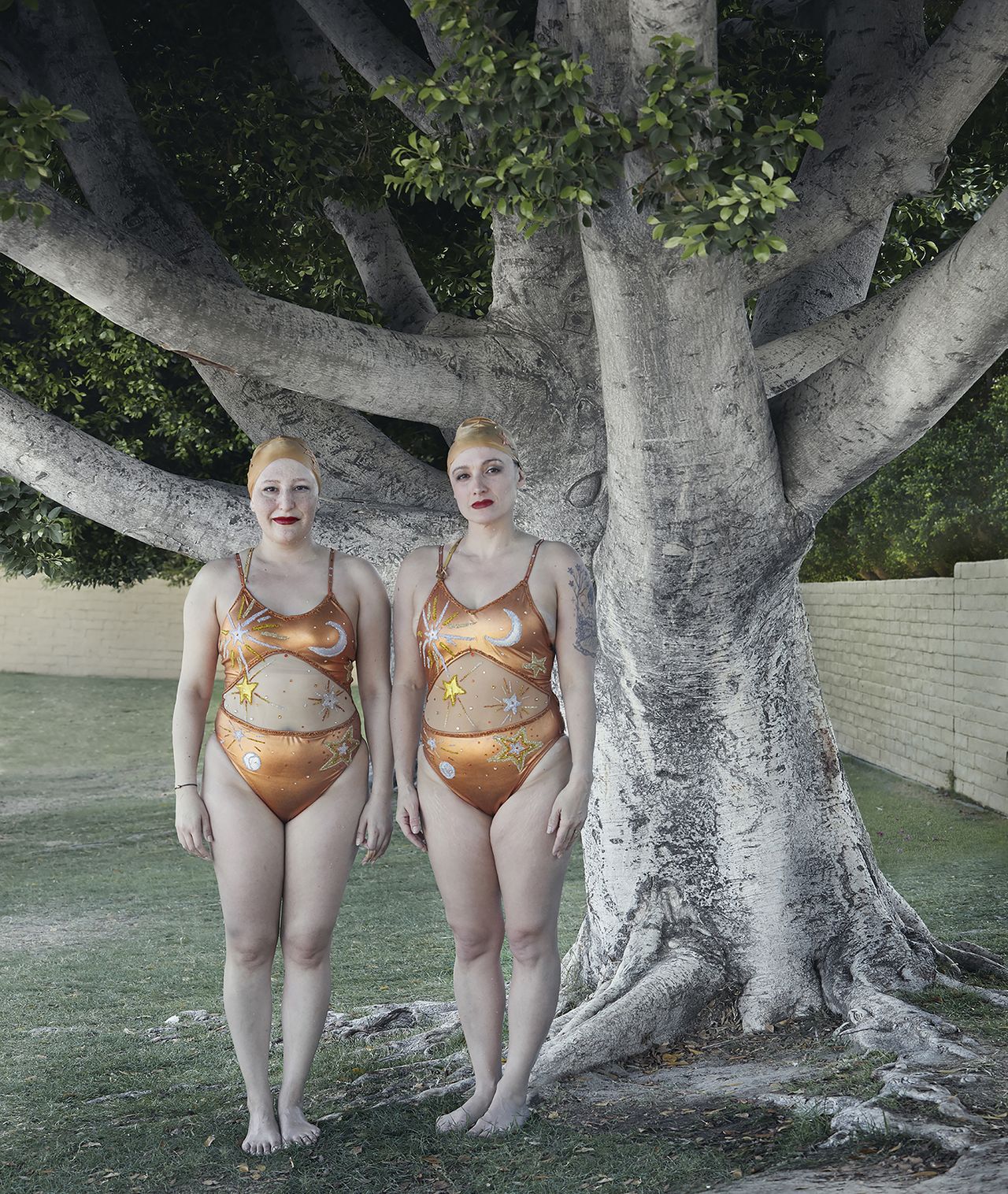 Two women in matching orange swimsuits with celestial designs stand barefoot in front of a large tree in a grassy area