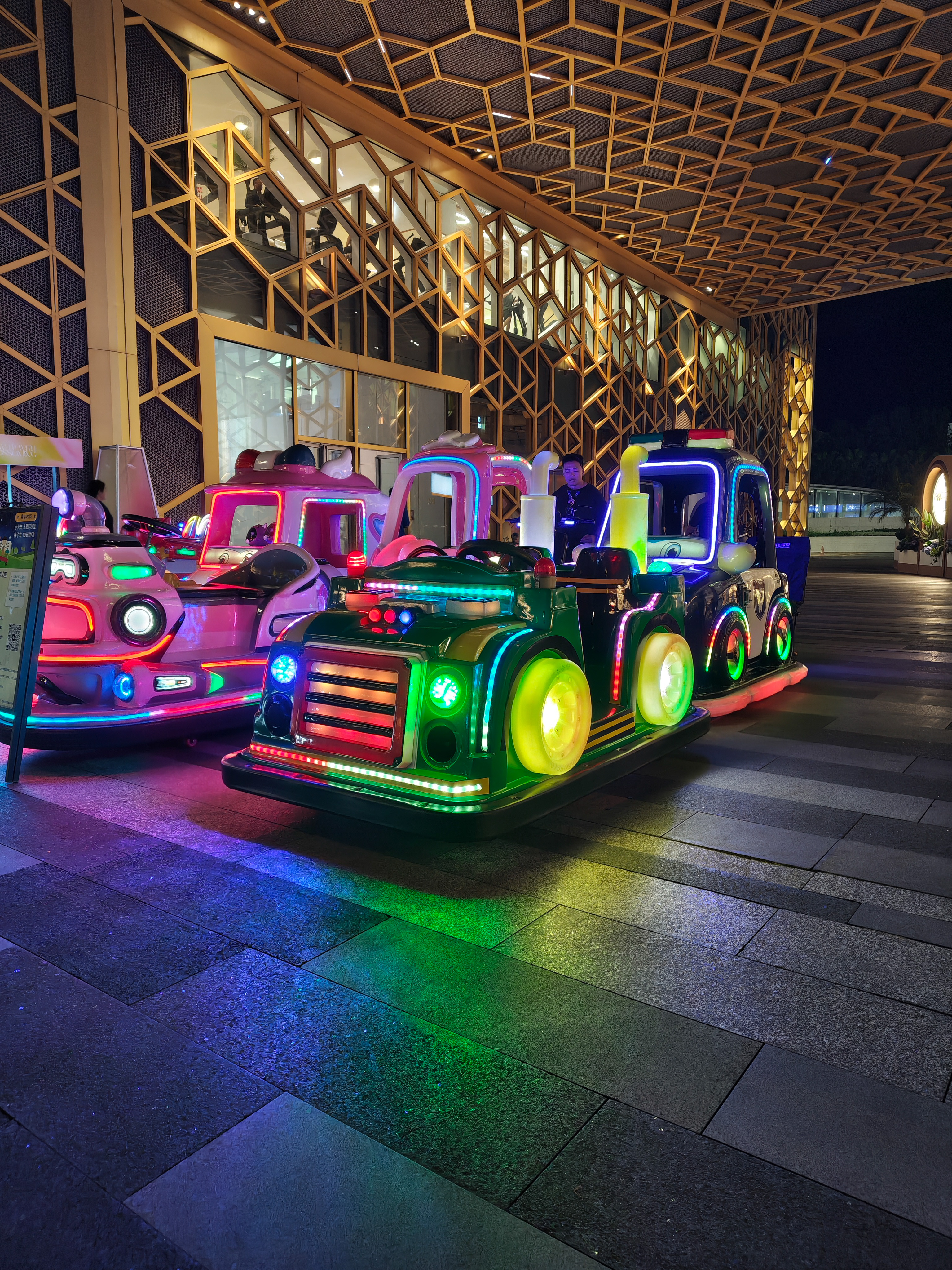 A night shot of several colorful children's toy train rides parked on a gray stone plaza. The trains are illuminated with bright neon green, blue, pink, and yellow lights. In the background, a building features a complex geometric gold lattice design over its windows.