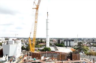 side view of a yellow crane hoisting a large white solid rocket motor into place in an unfinished building.