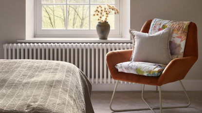 White radiator below a window. And orange chair sits next to it with a double bed just in front.