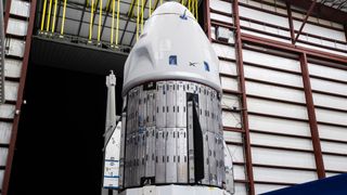 a white spacex dragon capsule is seen inside a large hangar near a door that opens onto darkness.
