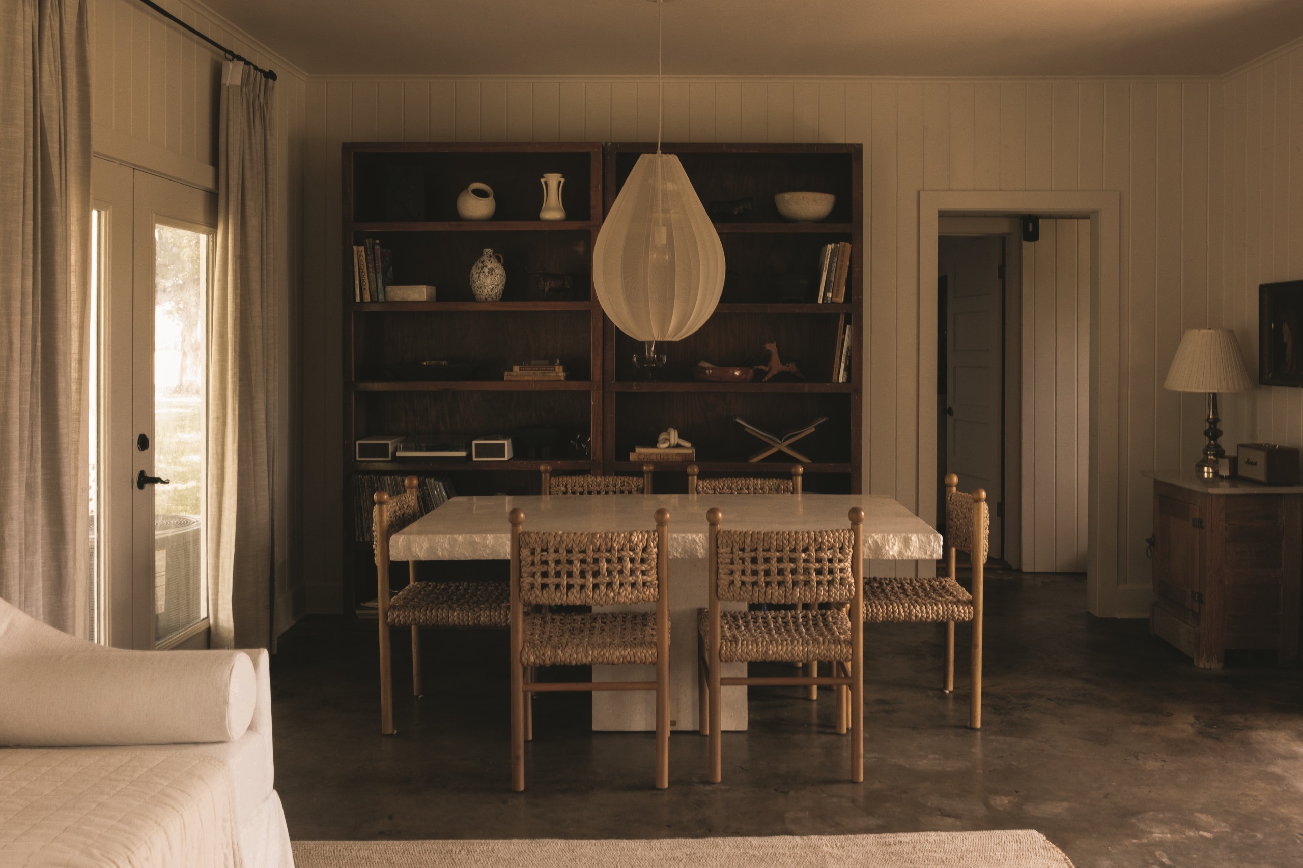 modern room with bookcase, pendant light, panelled walls with polished floors, travertine dining table and woven rattan chairs