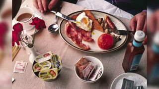 Close-up colour photograph of hands hold- ing a knife and fork and a full English breakfast with a cup of a hot beverage on the table, alongside fruit preservatives, sugar sachets, butter and a bottle of sauce.