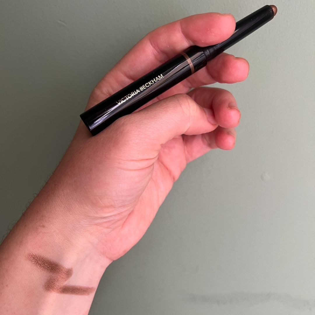 a hand on a green background holding Victoria Beckham EyeWear Longwear Eyeshadow with a swatch of the eyeshadow on the wrist 