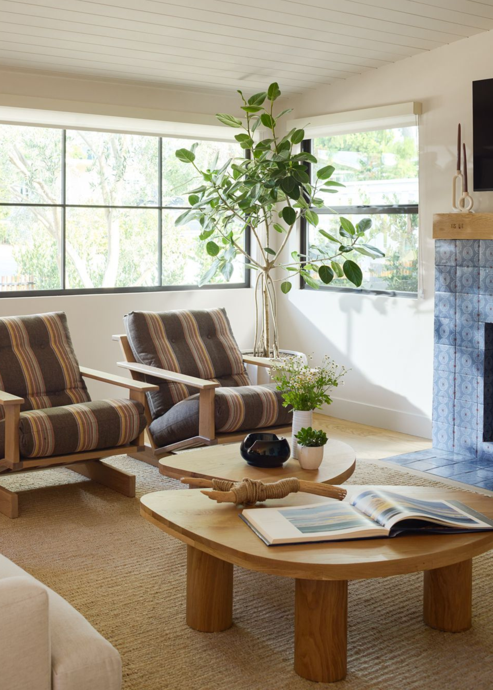 A living space with two irregular shaped coffee tables, one styled with an open book and a handmade slingshot, the other with a decorative bowl, a small plant and a vase of fresh flowers with two striped armchairs facing the tables next to a blue tiled fireplace with a large plant in the corner of the room
