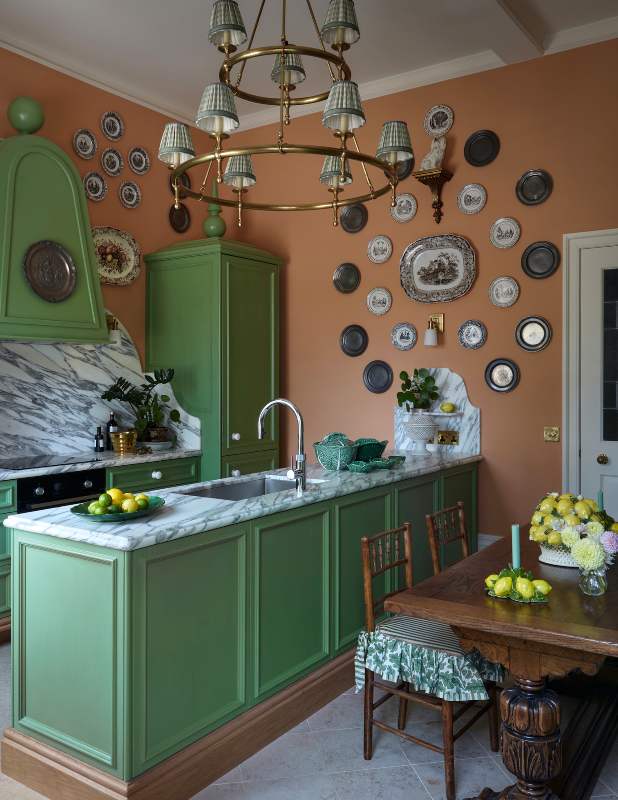 Green galley kitchen with a marble-topped sink peninsula beside a wooden dining table, peach walls displaying decorative plates and a brass chandelier with shaded lights.