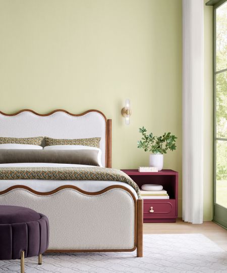 Bedroom painted in Sherwin-Williams' Celery, a wooden bed with scalloped headboard and end board, floral quilt on top, a purple stool, and a dark pink side table with a potted plant on top.