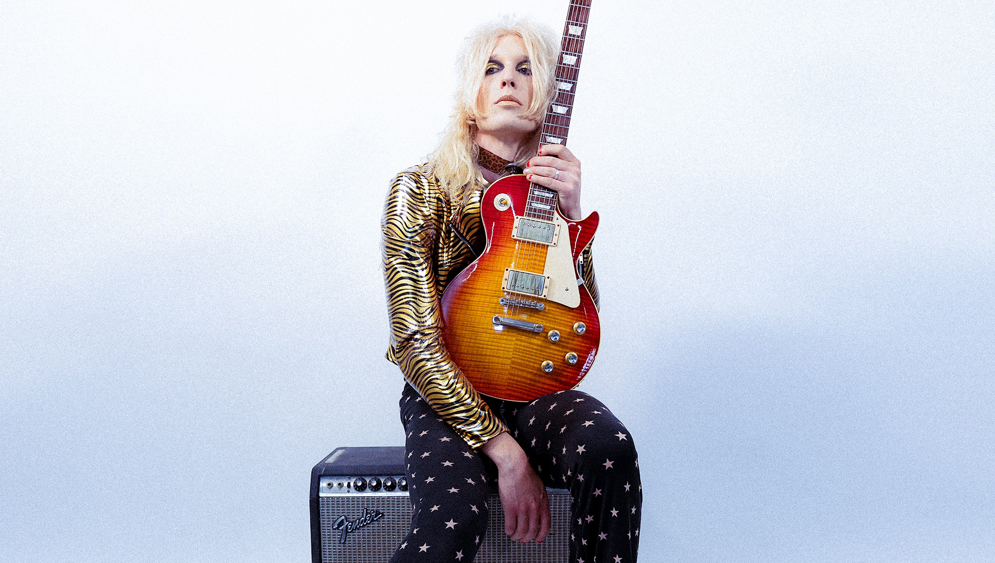 A photo of Alice Cooper band guitarist Gyasi holding a Gibson Les Paul guitar while sitting on an unidentifiable Fender combo