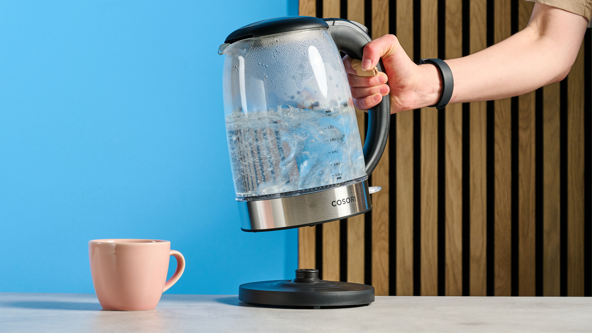 Cosori Electric Glass Kettle being held by a mug photographed in front of a blue background.