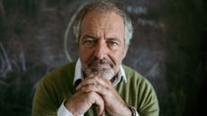 A mature, handsome man sits at home, hand on his chin, against a blackboard. He looks thoughtful.