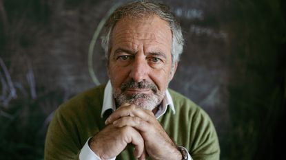 A mature, handsome man sits at home, hand on his chin, against a blackboard. He looks thoughtful.
