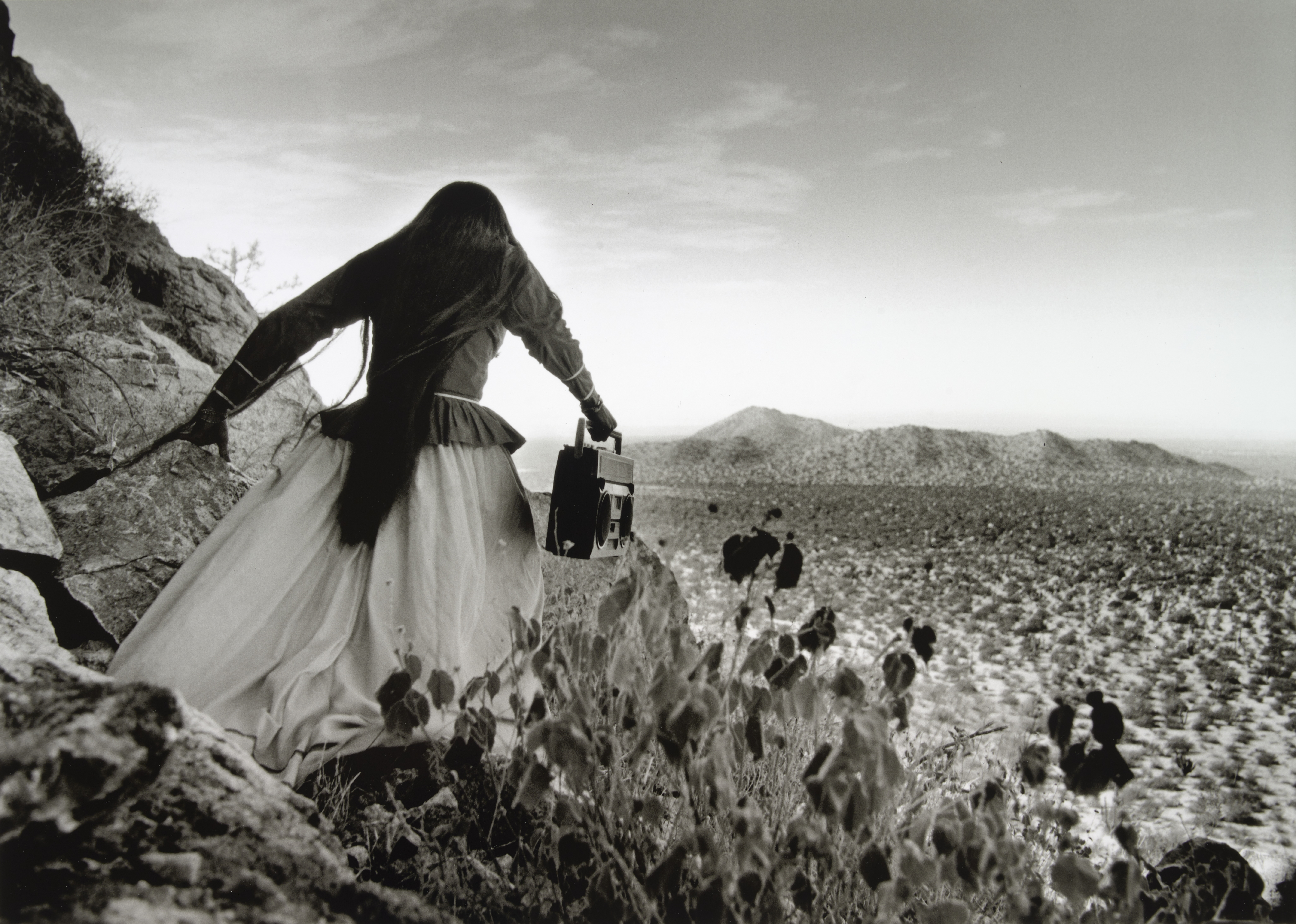 A woman in a long, wind-blown dress and dark flowing hair carries a boombox as she climbs a rocky desert hillside, seen from behind.