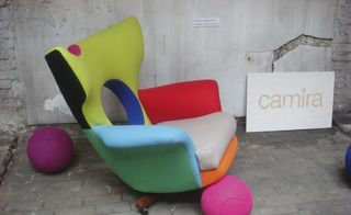 Grey brick floor, white rustic walls, multicoloured armchair design, two purple basketballs on the floor, 'Camira' in gold letters on a white sign leant against the back wall