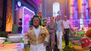 An Oompah Loompah in a yellow jacket escorts the contestants through Willy Wonka's Chocolate factory. The room is drenched in purple and pink hues.