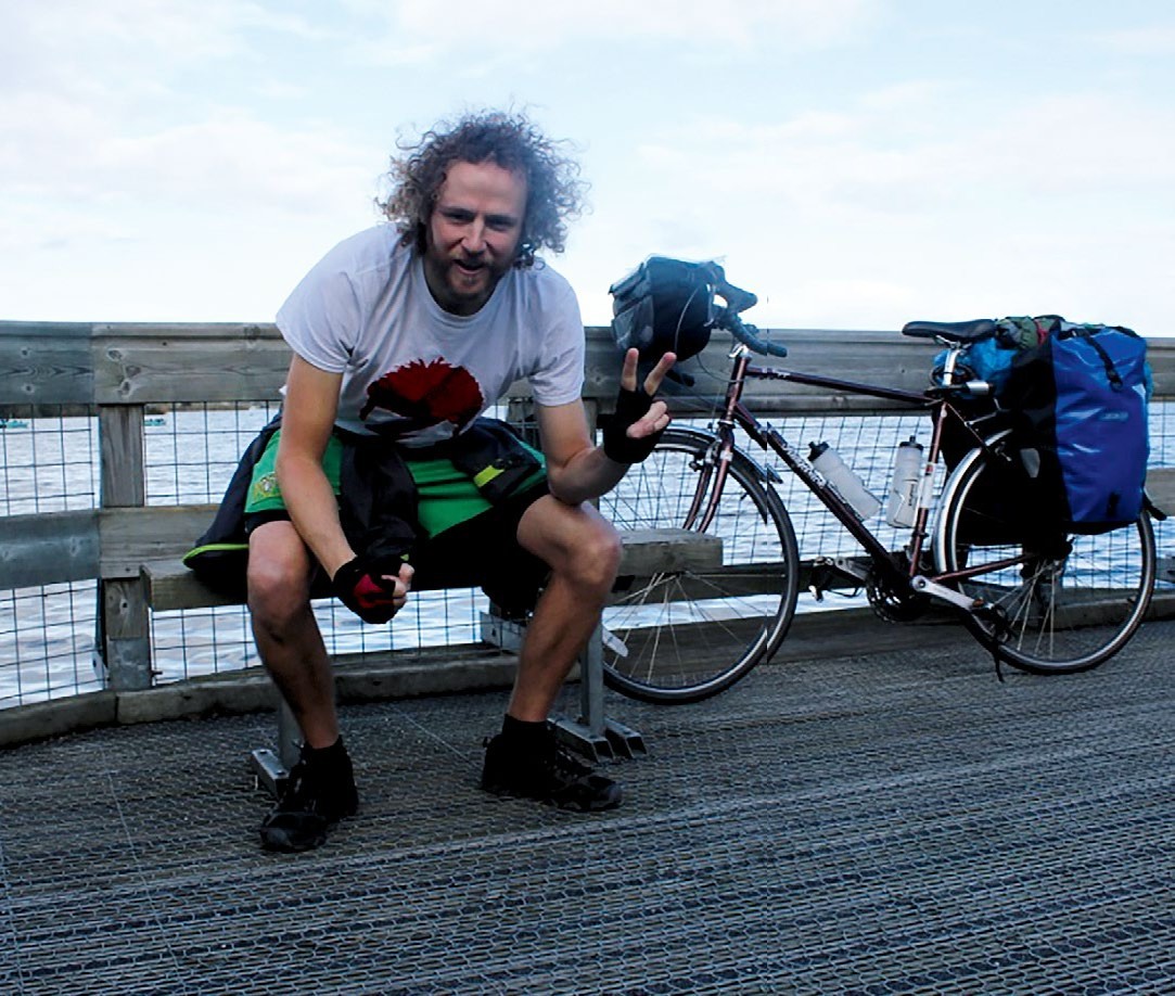 James Briggs sitting on a bench with wild hair, his bike beside him