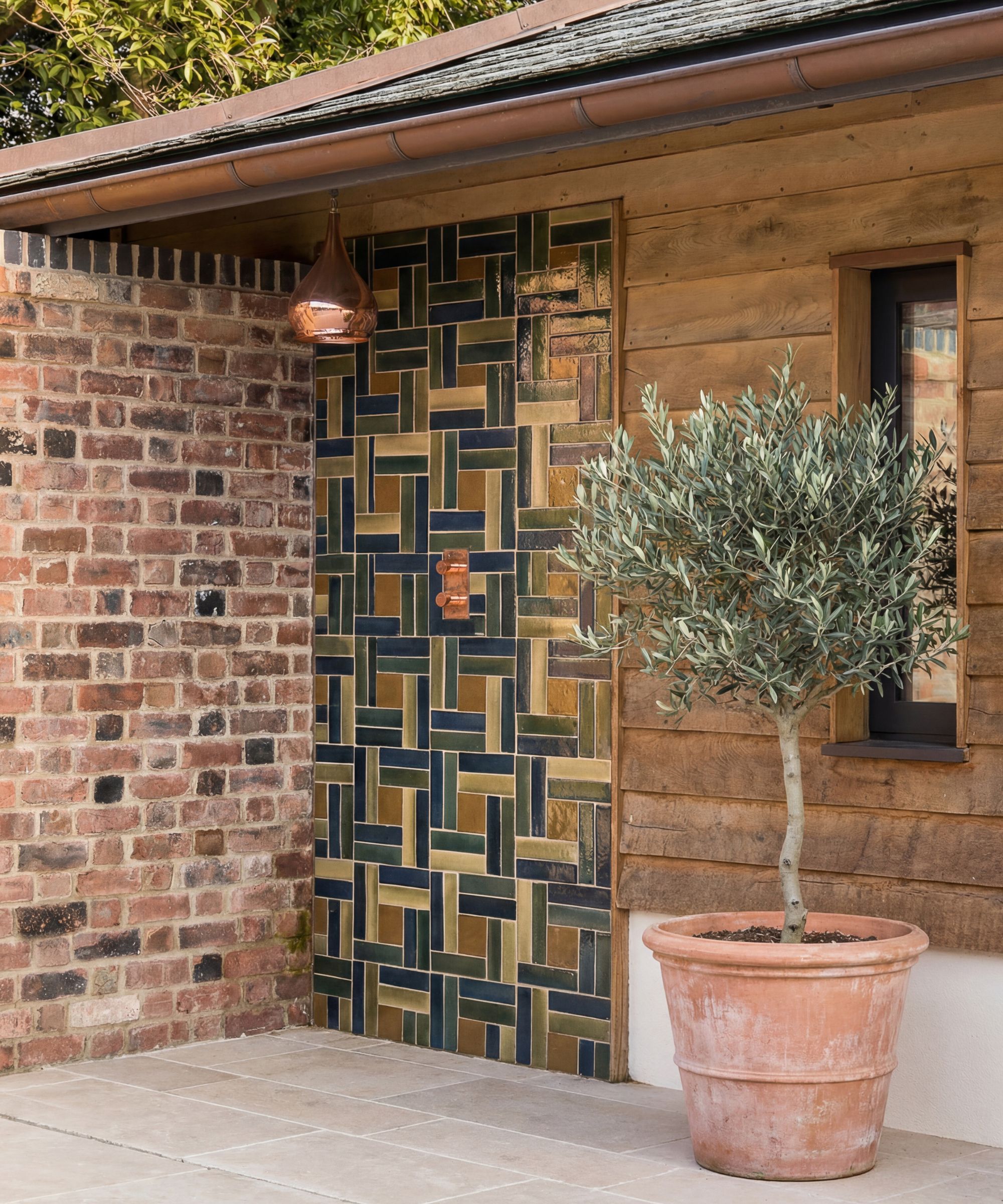 A copper pipe outdoor shower on a multicoloured glazed brick-pattern tiled panel on a timber-clad outbuilding, with a potted olive tree alongside.