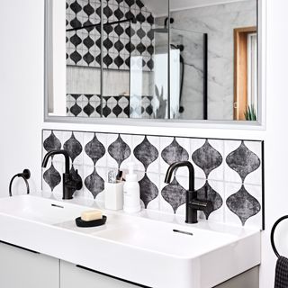 a bathroom basin with a black and white patterned tile splashback and black taps, a rectangular mirror, and black hand towels