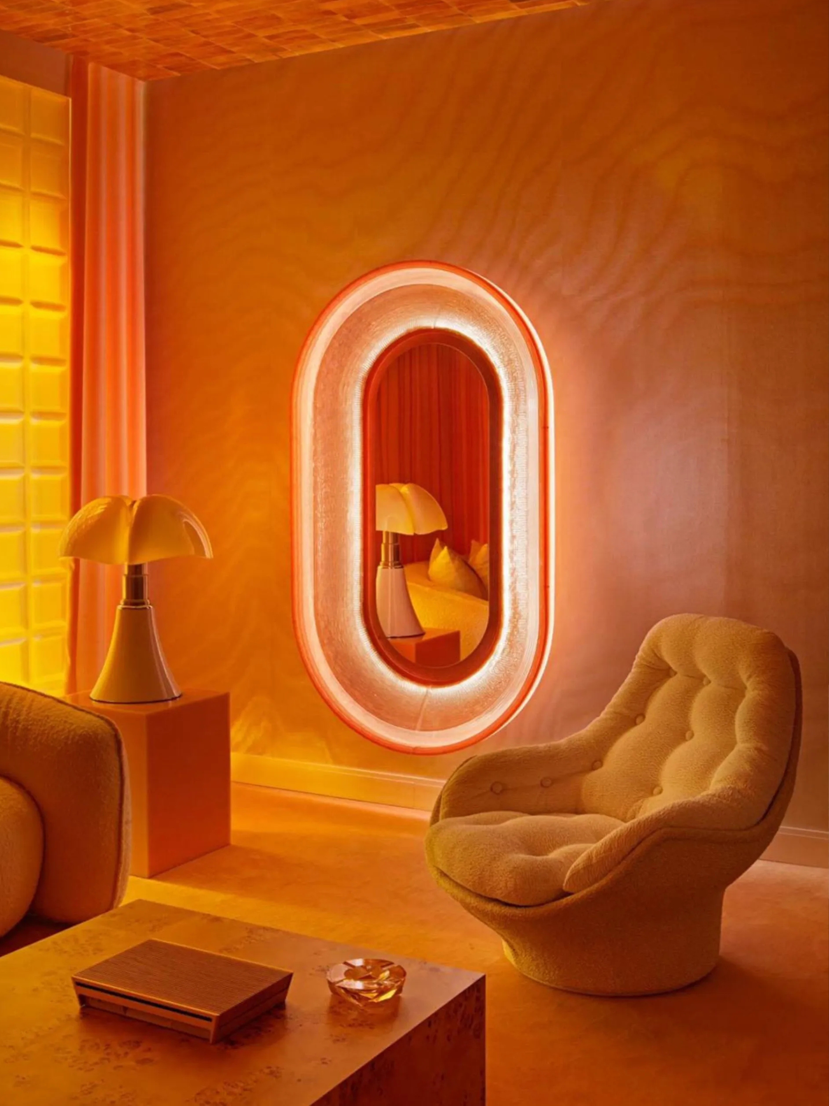 Image of a space-age-designed room that is color-drenched in orange. There is a bucket accent chair and a glowing mirror hanging on the wall.