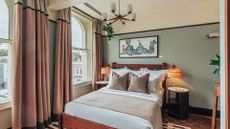 A hotel room with thick two-toned curtains, a large bed with white and tufted pillows, fringe lamps, marble nightstands, and a framed painting