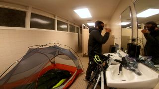 A man in black hoodie brushes his teeth in a public bathroom sink. Behind him is a single-person tent with a sleeping bag. Leaning against the sink is a silver bike.
